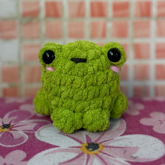 Green plush toy frog on a pink floral surface with a tiled wall background