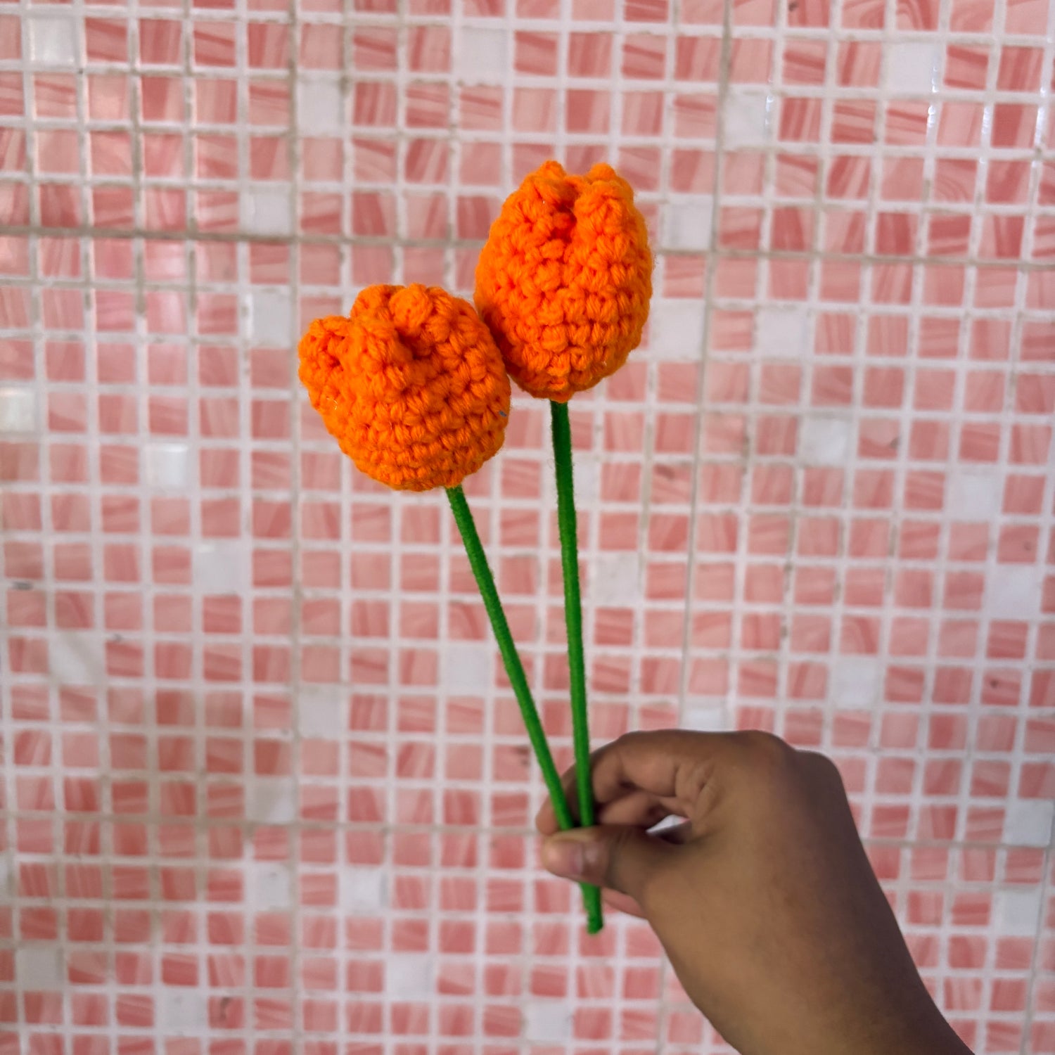 Hand holding two orange crocheted tulips against a pink tiled wall.
