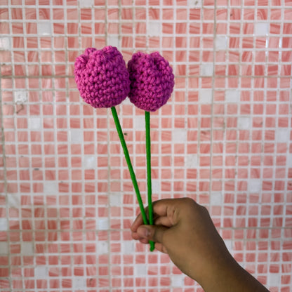Hand holding two purple crocheted flowers against a pink tiled wall.
