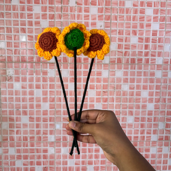 Hand holding three crocheted sunflower flowers against a pink tiled wall.