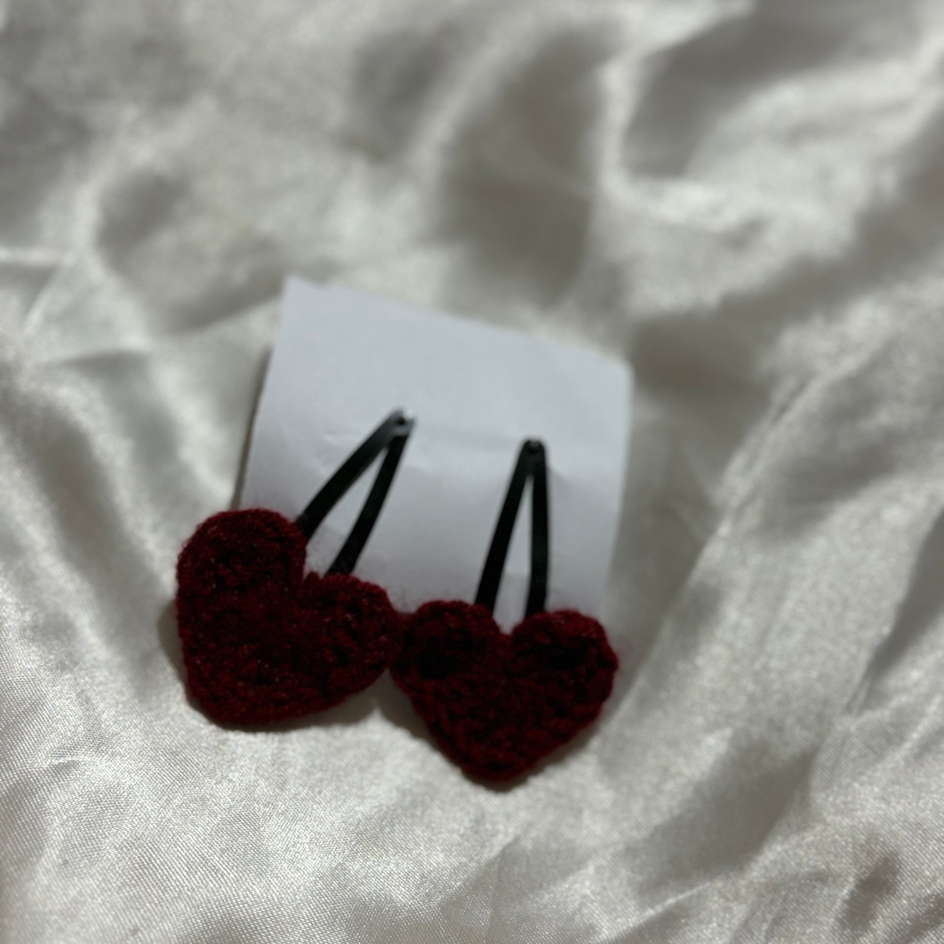 Two maroon heart-shaped hair clips on a white paper over a textured white fabric background.