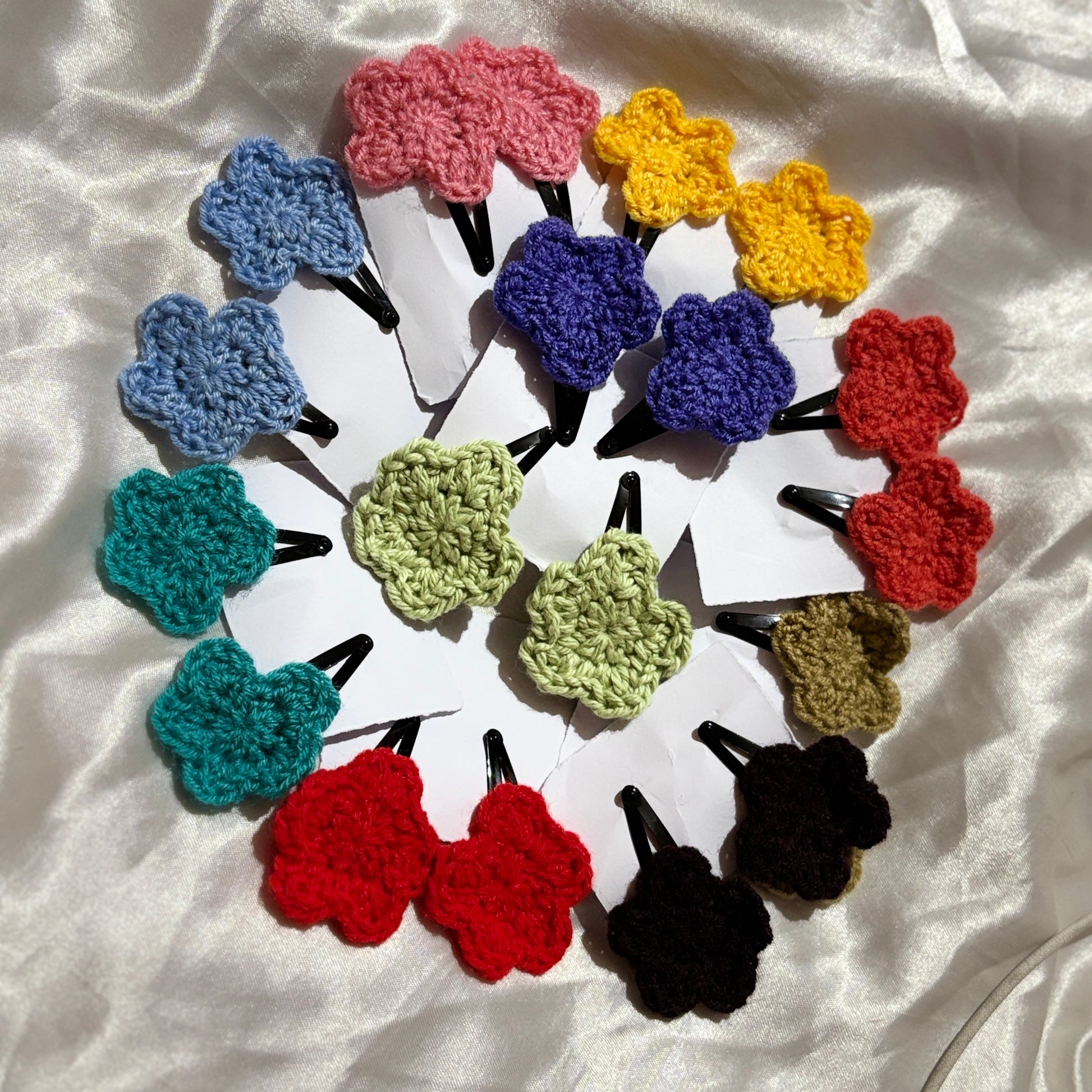Colorful crocheted hair clips arranged in a circular pattern on a white fabric background.