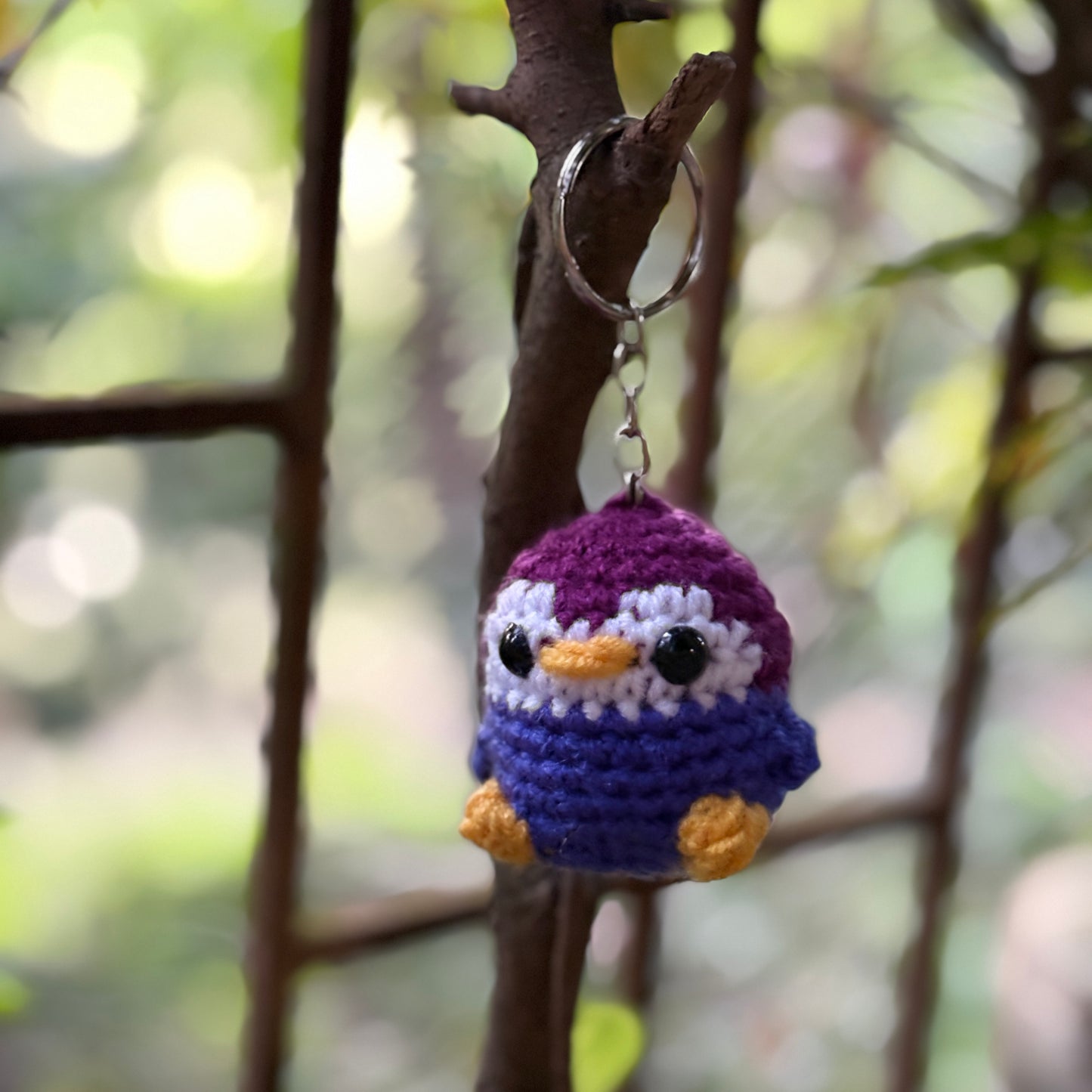 Crocheted penguin keychain hanging on a branch with a blurred natural background