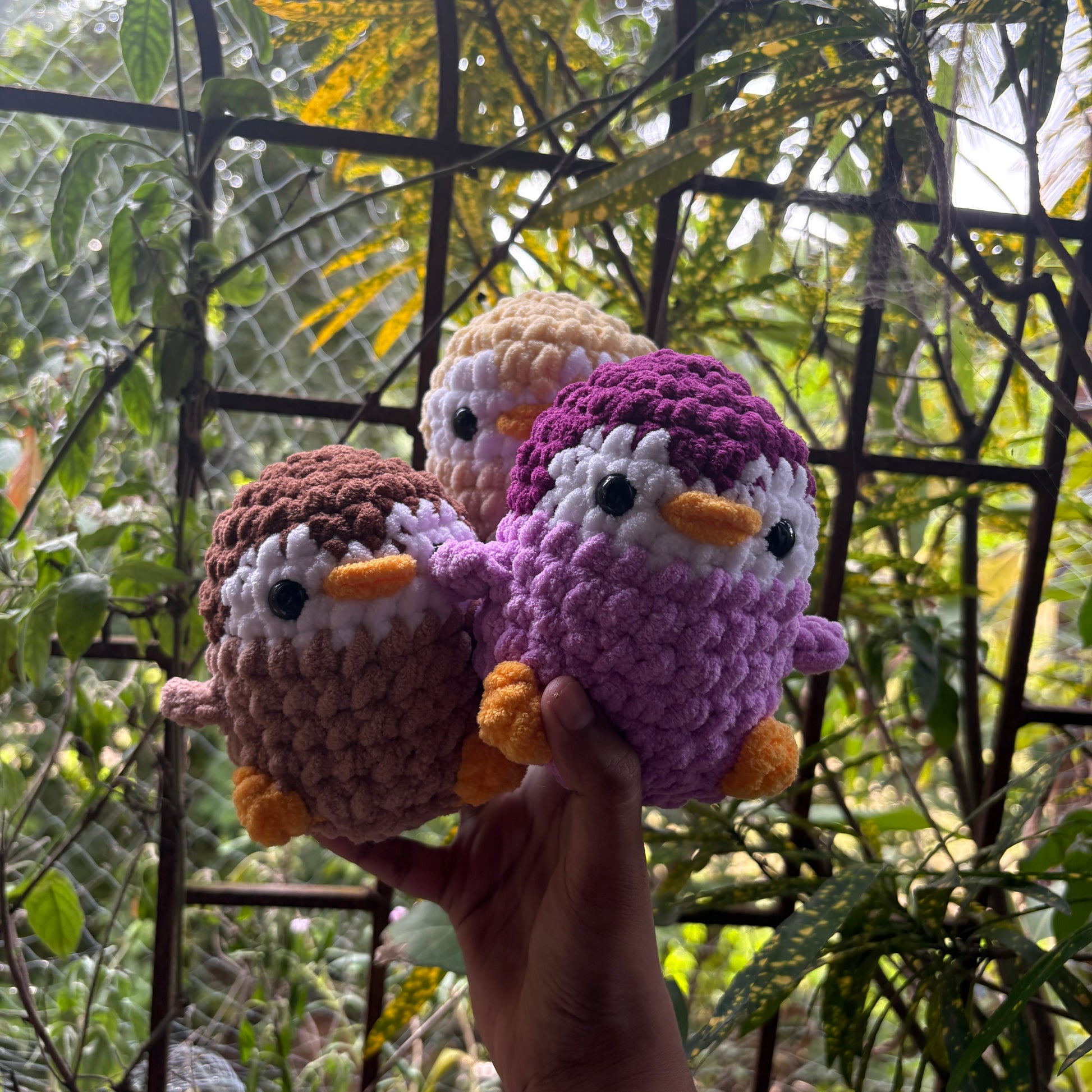 Three crocheted penguin toys held in front of a natural background with greenery.