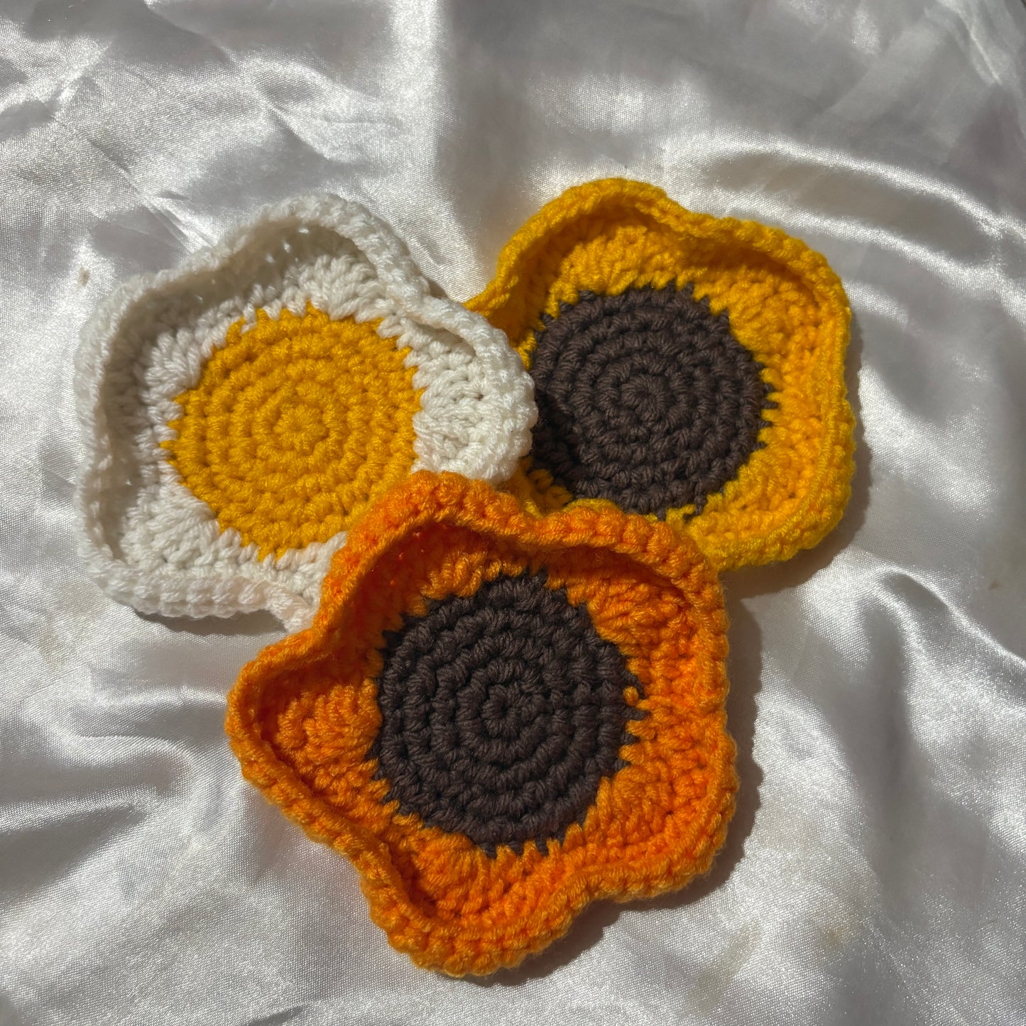 Three crocheted sunflower coasters in orange, yellow, and white on a white fabric background.