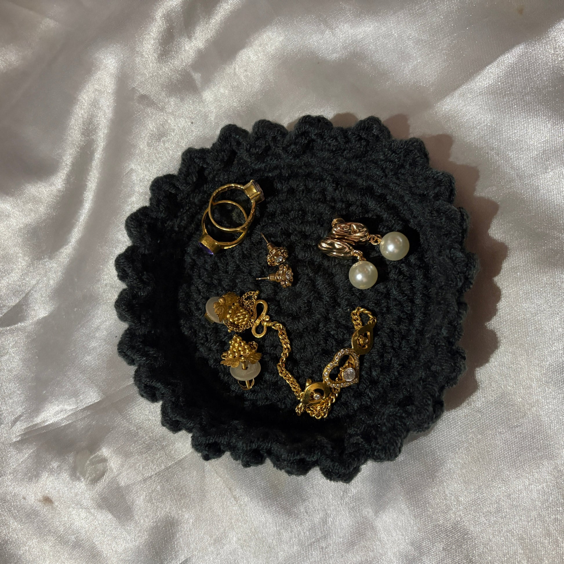 Black crochet dish with gold and pearl jewelry on a white fabric background