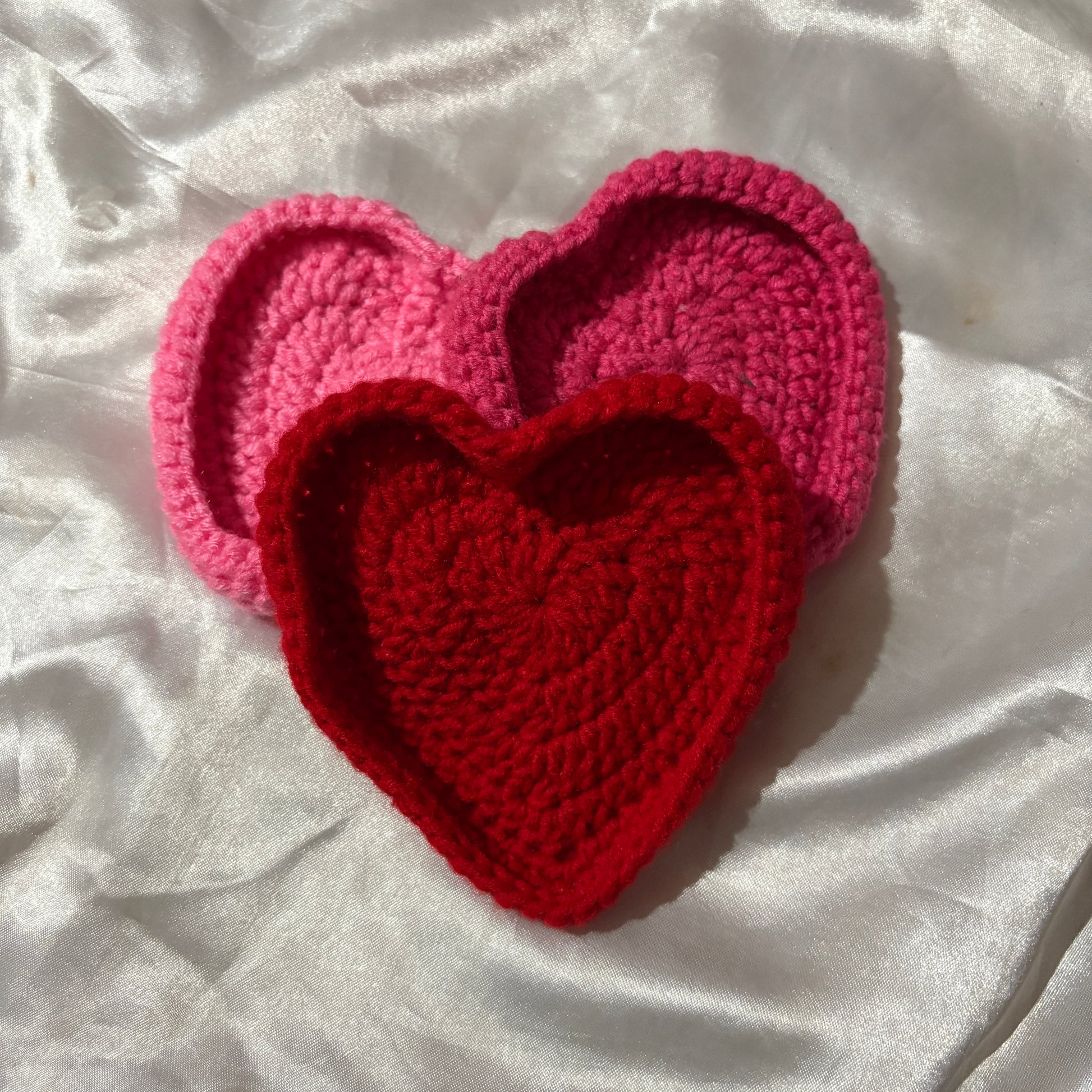 Two crocheted heart-shaped coasters, one red and one pink, on a white fabric background.