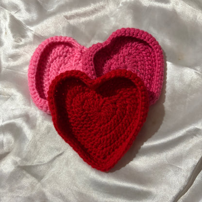 Two crocheted heart-shaped coasters, one red and one pink, on a white fabric background.