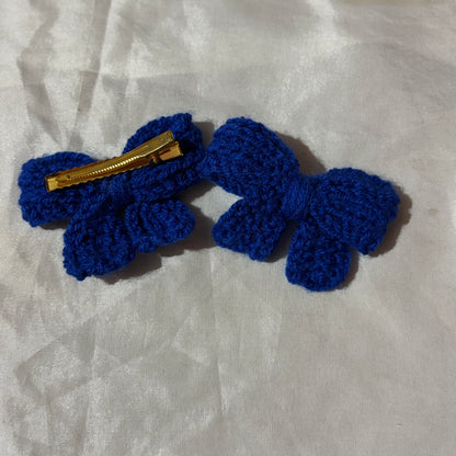 Blue crocheted bow clips with a gold clip on a light gray fabric background