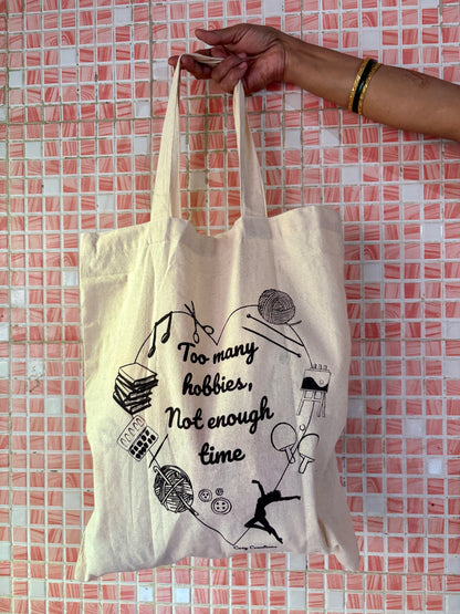 Tote bag with text and graphics held by a hand against a tiled wall background - too many hobbies not enough time