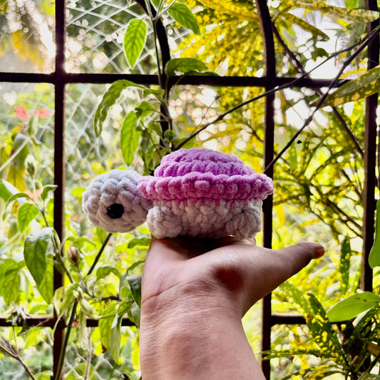 Hand holding a small crocheted turtle with a purple shell against a natural background
