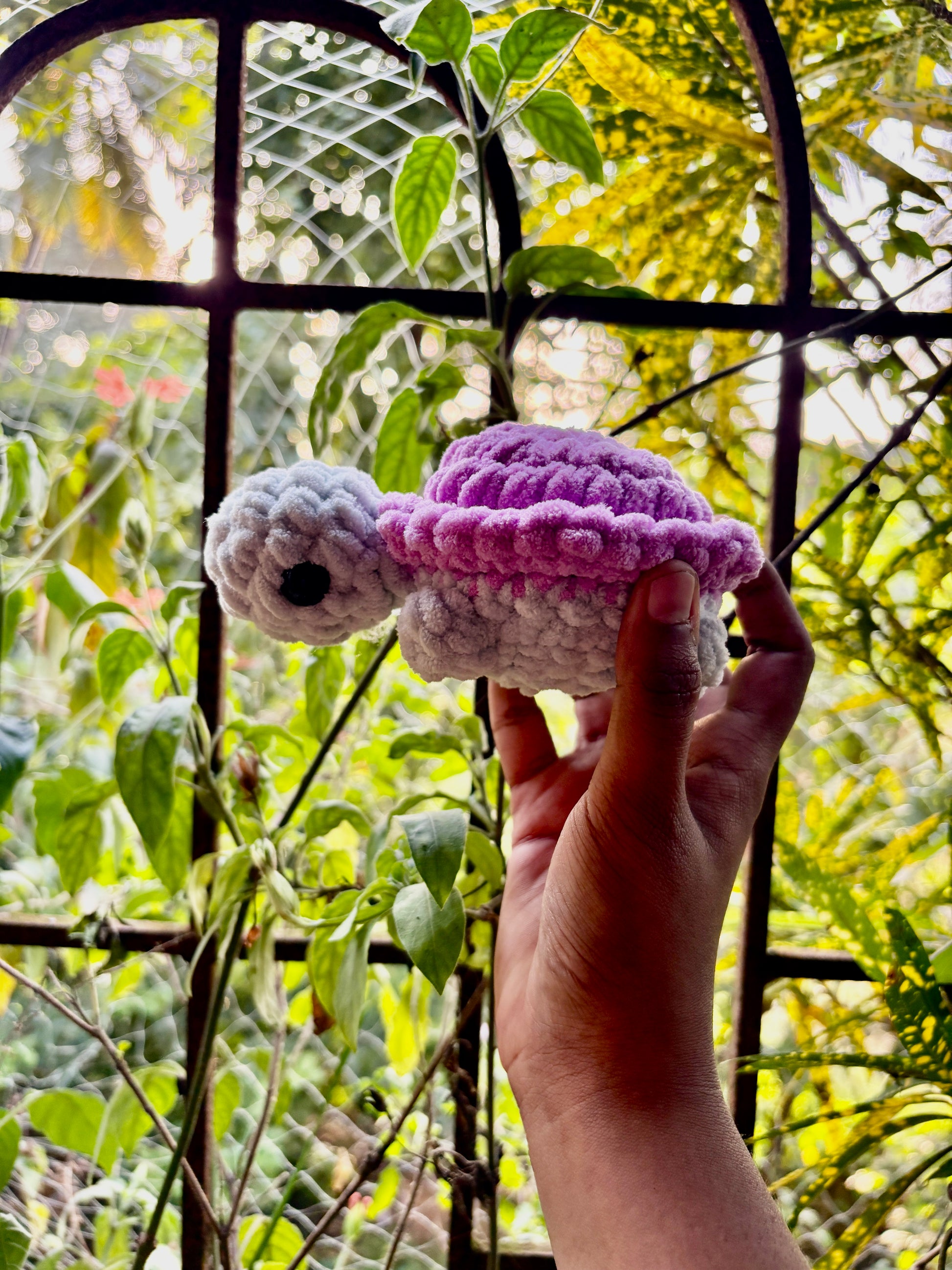 Hand holding a small crocheted turtle with a purple shell against a natural background