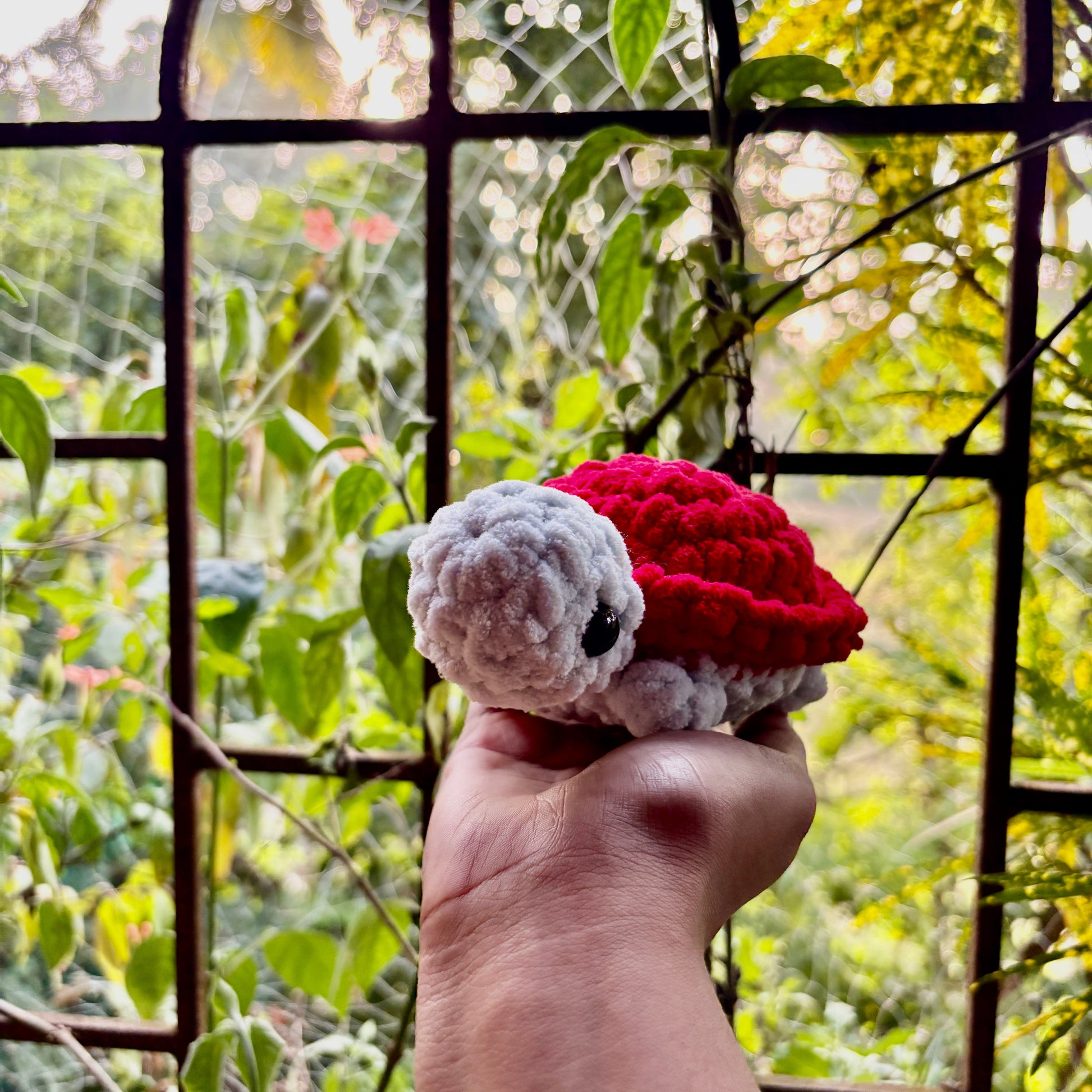 Hand holding a red crochet turtle toy in front of a window with greenery outside