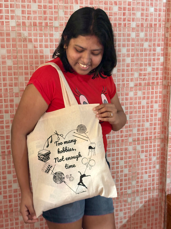 Person holding a tote bag with text and graphics in front of a window with plants. too many hobbies not enough time